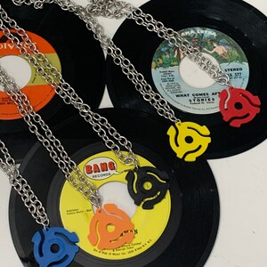 May include: A collection of vinyl record pendant necklaces in blue, orange, black, yellow, and red. Each pendant is a 45 RPM record adapter on a silver chain. The necklaces are displayed on top of vinyl records.