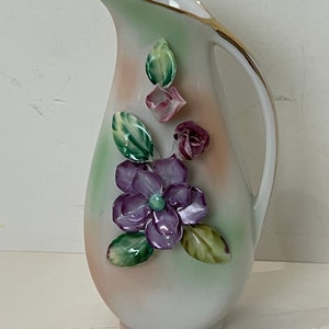 May include: A white ceramic pitcher with a gold rim and handle. The pitcher is decorated with raised purple and pink flowers and green leaves. The background has a soft gradient of peach and green.