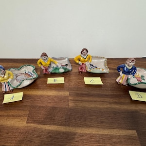 May include: Four ceramic trinket dishes, each featuring a hand-painted figure attached to the side. The figures wear yellow or blue tops and colorful skirts. Each dish has a unique painted design inside, labeled with yellow paper tags A, B, C, and D.