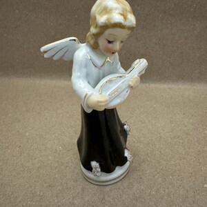 May include: A ceramic angel figurine with blonde hair, white wings, and a black dress. The angel is playing a white and gold instrument. The figurine has gold accents and is a decorative item.