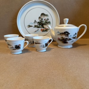 May include: A white porcelain tea set with a landscape design. The set includes a teapot, four teacups, and a serving tray. The landscape design features mountains, trees, and a river, with gold trim.