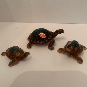 May include: Three ceramic turtle figurines. The turtles have brown bodies and dark green shells. Each turtle has a sticker that reads "Bone China Japan". The turtles are arranged on a white surface.