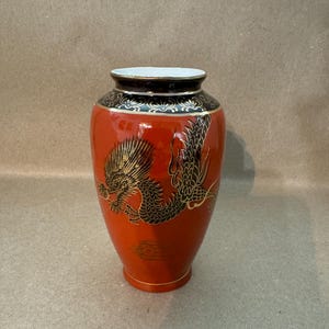May include: A red ceramic vase with a black and gold dragon design. The vase has a white interior and a black band with gold accents at the top. The dragon is detailed with gold and black, adding a touch of elegance.