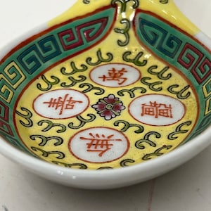 May include: A close-up of a small, decorative ceramic spoon with a yellow interior and white exterior. The spoon features a colorful design with red, green, and blue geometric patterns and Chinese characters. A small floral design is in the center.