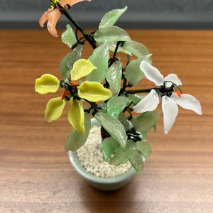 May include: A miniature glass bonsai tree with a light green ceramic pot. The tree features translucent green and yellow leaves, with delicate peach and white flowers. The trunk is dark, and the pot is filled with small white stones.