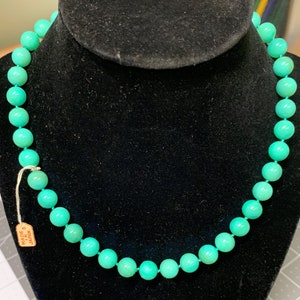 May include: A turquoise beaded necklace displayed on a black velvet bust. The necklace features round, evenly sized beads strung together. A small tag reading "Made in Japan" is attached. The beads are a vibrant, solid color.