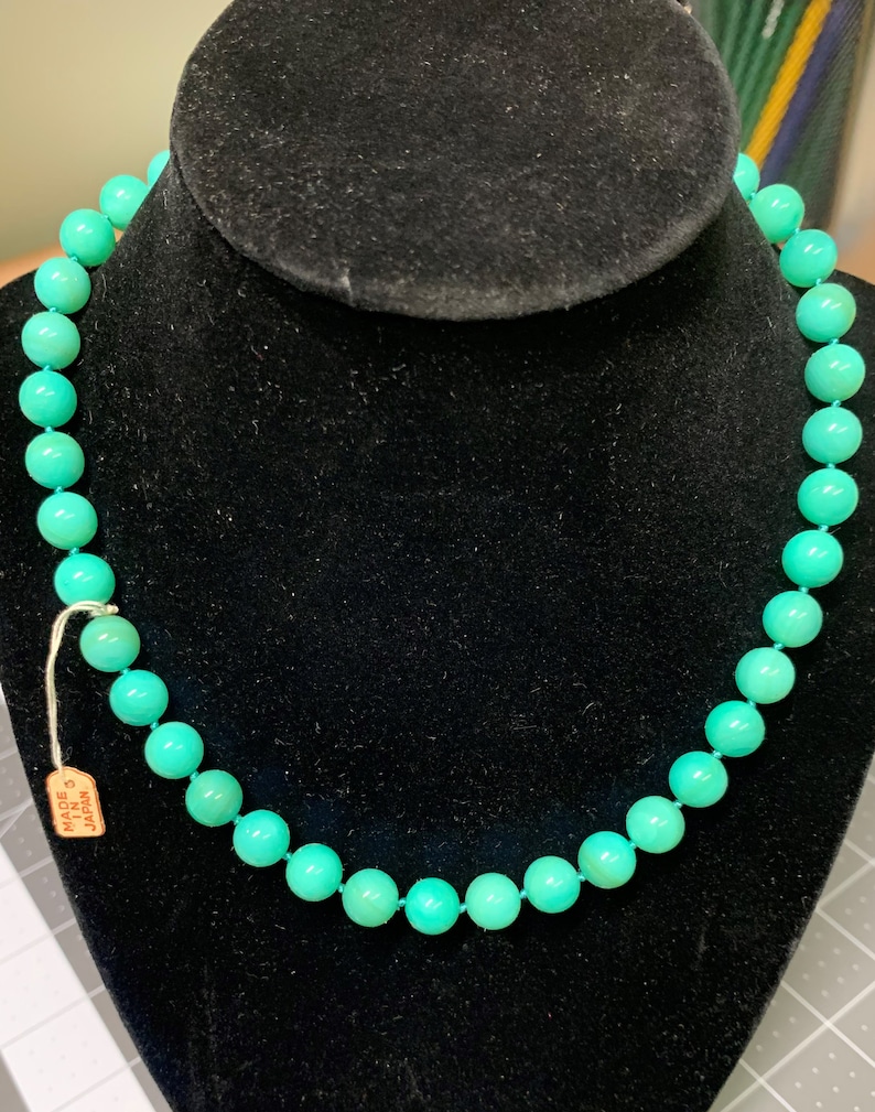May include: A turquoise beaded necklace displayed on a black velvet bust. The necklace features round, evenly sized beads strung together. A small tag reading "Made in Japan" is attached. The beads are a vibrant, solid color.