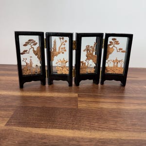 May include: A four-panel folding screen featuring black frames and intricate light brown cork carvings of landscapes, including trees, pagodas, and rock formations, connected by small gold-colored metal hinges.