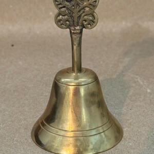 May include: A small, antique brass bell with a decorative floral design on the handle. The bell has a classic shape with a flared base and a detailed top. The brass has a warm, aged patina, giving it a vintage look.