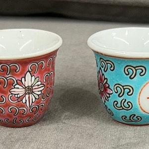 May include: Four small, decorative porcelain cups with white interiors and colorful exteriors. Two cups are red with white floral designs and Chinese characters. The other two are turquoise with Chinese characters and floral designs.