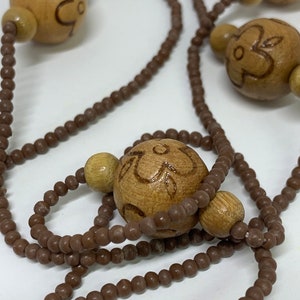 May include: A necklace featuring large, light brown wooden beads with etched floral designs, interspersed with smaller, round brown beads. The necklace has a natural, earthy aesthetic.