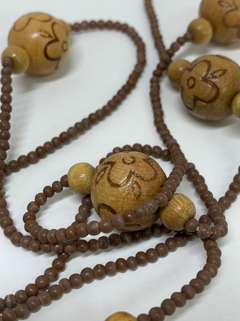 May include: A necklace featuring large, light brown wooden beads with etched floral designs, interspersed with smaller, round brown beads. The necklace has a natural, earthy aesthetic.