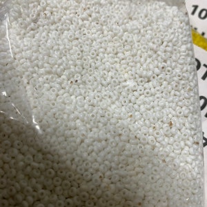 May include: A clear plastic bag filled with numerous small, white, round beads. The beads appear to be uniform in size and shape, with a slightly glossy finish. The beads are tightly packed together, filling the majority of the frame.