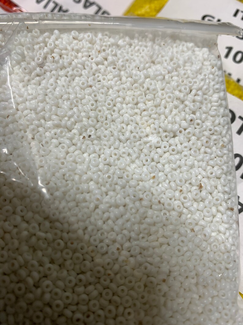 May include: A clear plastic bag filled with numerous small, white, round beads. The beads appear to be uniform in size and shape, with a slightly glossy finish. The beads are tightly packed together, filling the majority of the frame.