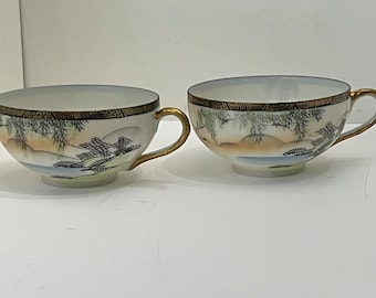 May include: Two white porcelain teacups with gold handles and rims. Each cup features a hand-painted landscape scene with trees, mountains, and water in shades of black, blue, and tan. The rims have a decorative black and gold border.
