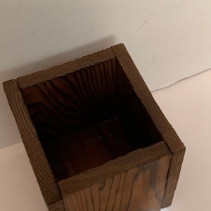 May include: A rectangular wooden box with a dark brown finish. The box has a textured, ridged border around the top edge. The interior of the box is a darker shade of brown. The wood grain is visible on the sides.