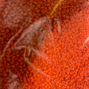 May include: Close-up of a bag filled with numerous small, orange seed beads. The beads are uniform in size and shape, with a hole running through the center of each. The beads are a vibrant, solid orange color.
