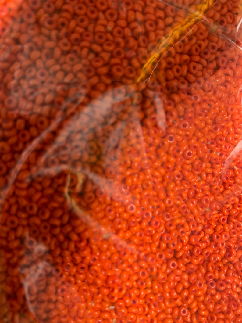 May include: Close-up of a bag filled with numerous small, orange seed beads. The beads are uniform in size and shape, with a hole running through the center of each. The beads are a vibrant, solid orange color.