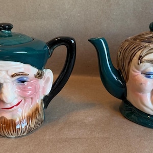 May include: Two ceramic teapots with human face designs. One teapot has a green hat and a beard, the other has brown hair and closed eyes. Both teapots have black handles and spouts, and are painted with pink and red accents.