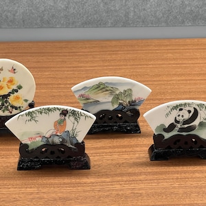 May include: Four decorative ceramic pieces on wooden stands. One is round with yellow roses, and the others are fan-shaped, each with a different Asian-inspired design: a landscape, a woman, and a panda. The stands are dark brown.