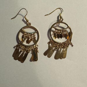 May include: A pair of gold-colored dangle earrings. Each earring features a circular design with smaller dangling elements within the circle and a fringe of flat, teardrop-shaped pendants. The earrings have a hook closure.