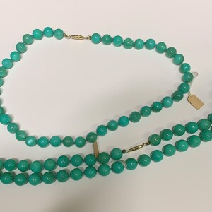 May include: Two turquoise beaded necklaces with gold clasps are displayed against a white background. The necklaces are composed of round, polished beads. One necklace is doubled, and both are connected with gold clasps.