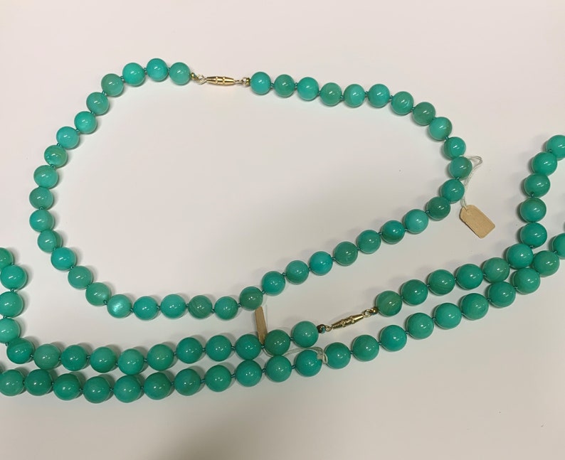 May include: Two turquoise beaded necklaces with gold clasps are displayed against a white background. The necklaces are composed of round, polished beads. One necklace is doubled, and both are connected with gold clasps.