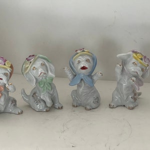 May include: Four ceramic dog figurines in various poses. Each dog is white with gold accents, wearing a bonnet or scarf with floral details and a bow. The dogs have red mouths and are in a playful, vintage style.