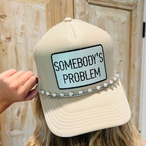 May include: Tan trucker hat with a mesh back and a curved brim. The front features a rectangular patch with the text "SOMEBODY'S PROBLEM" in black. A string of white beads decorates the brim.