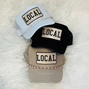 May include: Three trucker hats with a gold "LOCAL" patch and a pearl chain. The hats are white, black, and beige.
