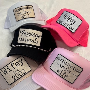 May include: A collection of trucker hats in white, black, and pink, each featuring a rectangular patch with text. The patches read "Participation Trophy Wife," "Marriage Material," and "Wifey," with one hat also stating "Est. 2002."