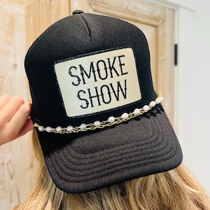 Puede incluir: Gorra de camionero negra con un parche blanco que dice "SMOKE SHOW" en letras negras. El sombrero presenta un detalle de cadena de perlas y oro en el borde. La persona que lleva el sombrero tiene el pelo rubio.
