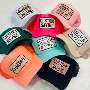 May include: A collection of trucker hats in various colors, including white, pink, green, orange, blue, and black. Each hat features a rectangular patch with the words "Custom Saying" in a bold, black font.