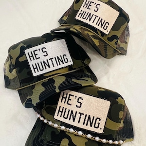 May include: Four camouflage trucker hats with the text "HE'S HUNTING" on a white patch. The hats have a mesh back and a curved brim. One hat has a pearl chain along the brim.
