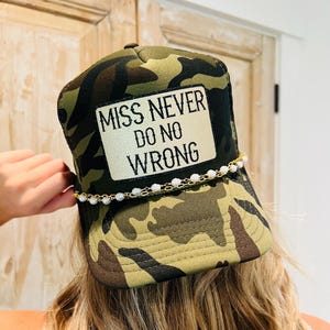 May include: Camouflage print baseball cap with the text "MISS NEVER DO NO WRONG" on a white patch. The cap features a gold chain with white pearls. The hat is a mix of green, brown, and black.