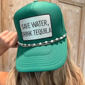 May include: A green trucker hat with a white patch that reads "SAVE WATER, DRINK TEQUILA" in black text. The hat has a pearl and silver chain detail along the brim.