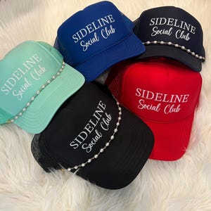 May include: Five trucker hats in mint green, royal blue, navy blue, black, and red. Each hat features the white text "SIDELINE Social Club" printed on the front. The mint green, navy blue, and black hats are adorned with a string of white pearls.