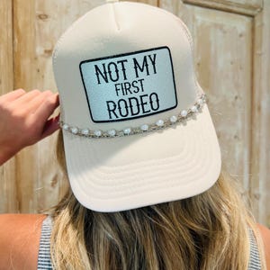 May include: Cream-colored trucker hat with the text "NOT MY FIRST RODEO" on a rectangular patch. The hat features a pearl and chain detail along the brim. The hat is worn by a person with blonde hair.
