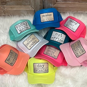 May include: A collection of trucker hats in various colors, including teal, pink, and yellow. Each hat features a rectangular patch with phrases like "Vacay Mode," "Sunshine and Seltzers," and "Day Drinking." The hats have mesh backs.
