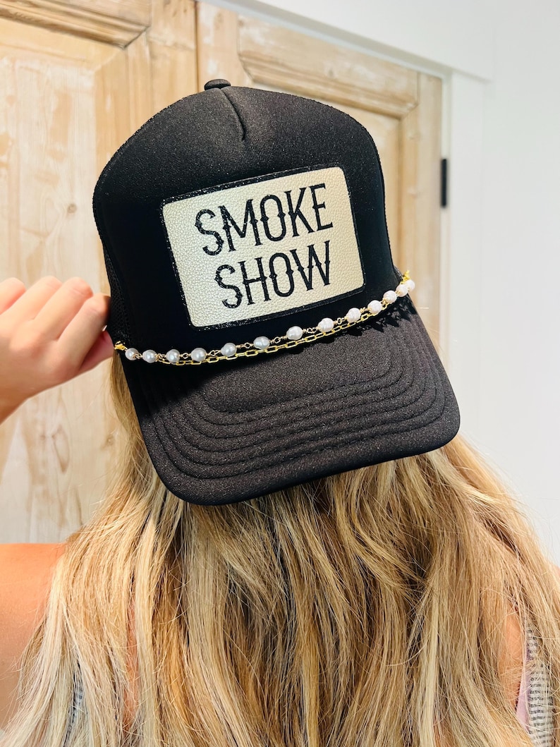 Puede incluir: Gorra de camionero negra con un parche blanco que dice "SMOKE SHOW" en letras negras. El sombrero presenta un detalle de cadena de perlas y oro en el borde. La persona que lleva el sombrero tiene el pelo rubio.