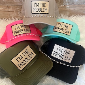 May include: Five trucker hats in beige, hot pink, mint green, olive green, and black. Each hat features a rectangular patch with the text I'M THE PROBLEM in black lettering. The hats are accented with a single strand of white faux pearls.