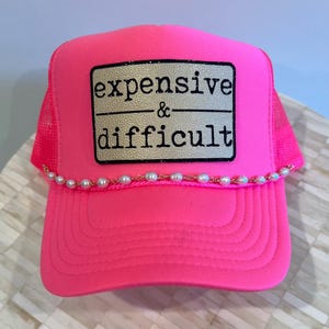 May include: Bright pink trucker hat with a pearl beaded band and a rectangular patch that reads "expensive & difficult" in black text. The hat has a mesh back and a curved brim.
