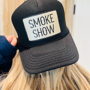 Puede incluir: Gorra de camionero negra con un parche blanco con el texto "SMOKE SHOW" en negro. La gorra tiene una visera curva y una parte trasera de malla. Un accesorio cl&aacute;sico.