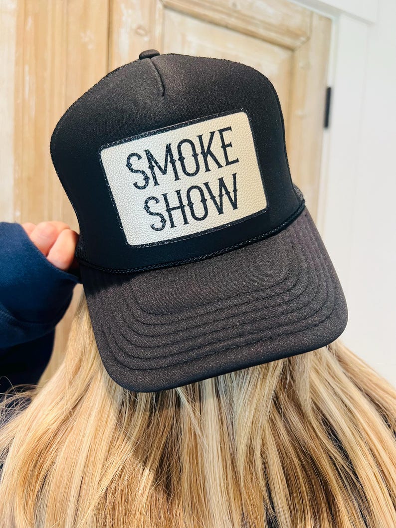 Puede incluir: Gorra de camionero negra con un parche blanco con el texto "SMOKE SHOW" en negro. La gorra tiene una visera curva y una parte trasera de malla. Un accesorio cl&aacute;sico.
