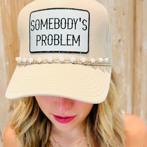Puede incluir: Gorra de camionero beige con el texto "SOMEBODY'S PROBLEM" en un parche blanco. La gorra tiene una visera curva y un adorno de cuentas. La gorra es de color neutro y tiene un dise&ntilde;o cl&aacute;sico de gorra de camionero.