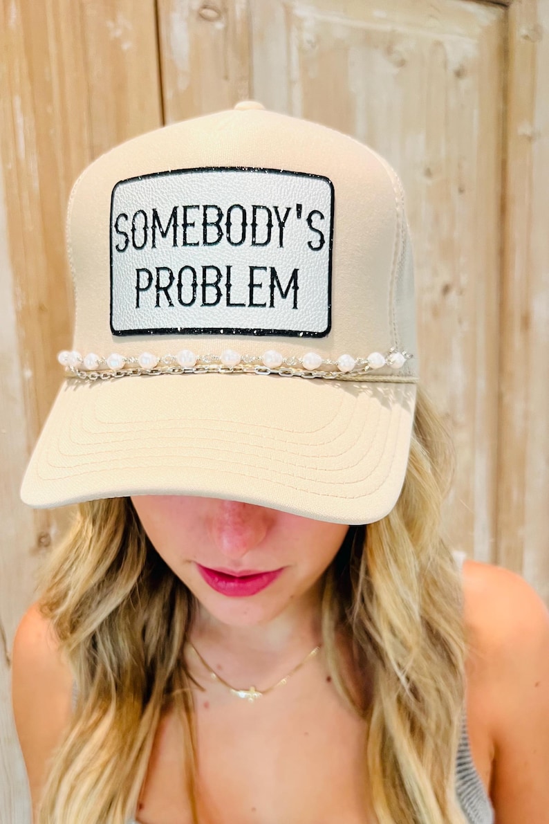 Puede incluir: Gorra de camionero beige con el texto "SOMEBODY'S PROBLEM" en un parche blanco. La gorra tiene una visera curva y un adorno de cuentas. La gorra es de color neutro y tiene un dise&ntilde;o cl&aacute;sico de gorra de camionero.