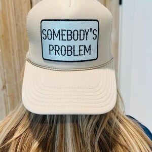 Puede incluir: Gorra de camionero beige con un parche rectangular que dice "SOMEBODY'S PROBLEM" en texto negro. La gorra tiene una visera curva y una parte trasera de malla.