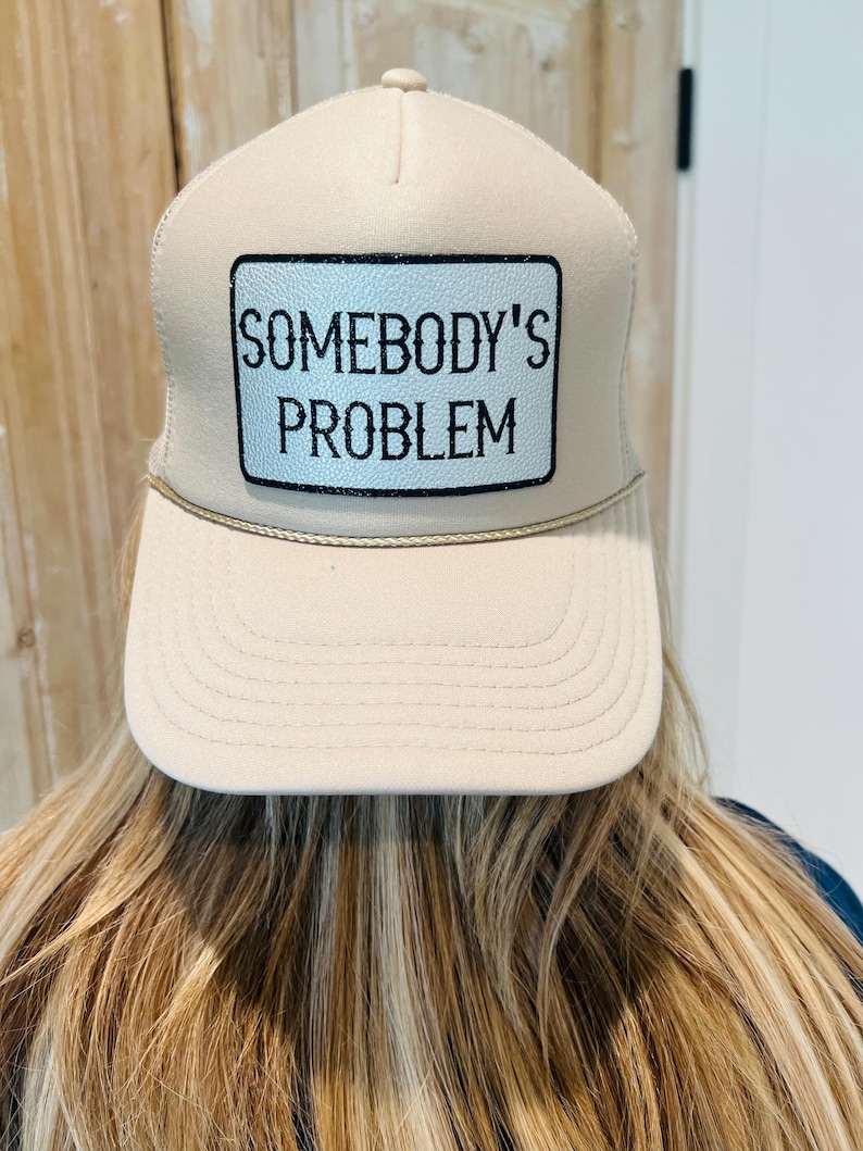 Puede incluir: Gorra de camionero beige con un parche rectangular que dice "SOMEBODY'S PROBLEM" en texto negro. La gorra tiene una visera curva y una parte trasera de malla.