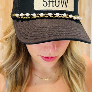 Puede incluir: Gorra de camionero negra con el texto "SMOKE SHOW" en un parche beige. La gorra presenta un detalle de cadena de perlas y oro en el borde. Un accesorio elegante para uso casual.