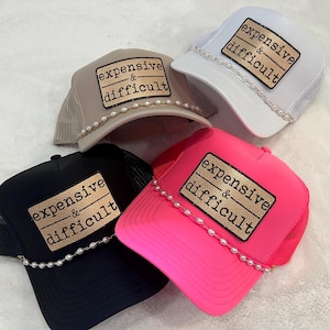 May include: Four trucker hats in black, white, beige, and neon pink. Each hat features a rectangular gold patch with the text "expensive & difficult" in black lettering and a decorative string of white faux pearls across the brim base.